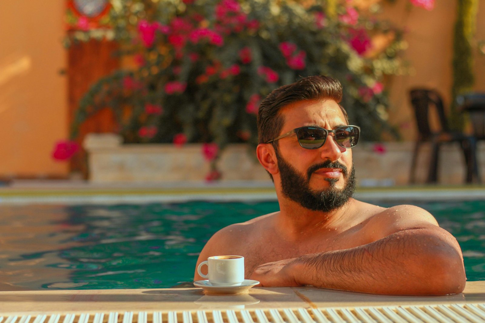 Man enjoying a sunny poolside moment with coffee, wearing sunglasses.